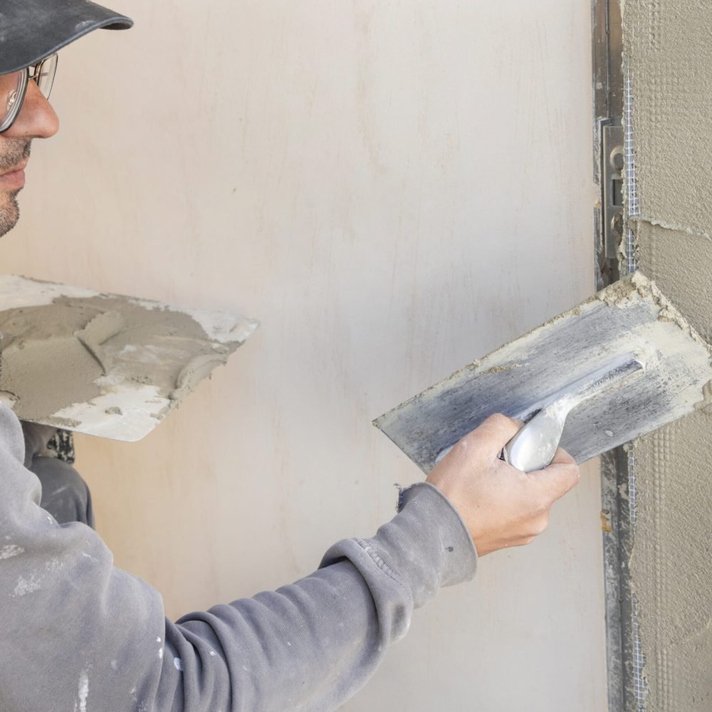Craftsman uses a float trowel and cement to plaster a house wall. Young men with glasses in gray work clothing during construction or renovation process