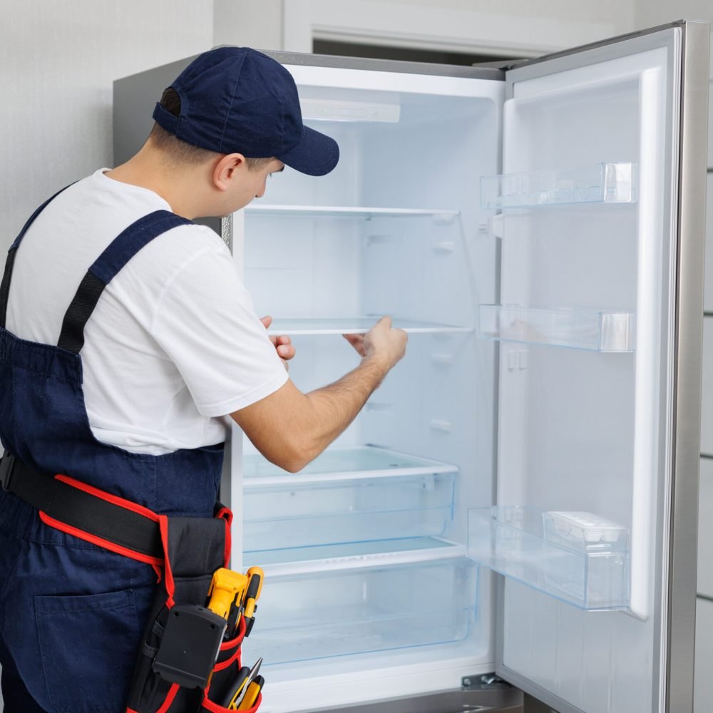 Man in a uniform with tools is repairing a refrigerator in the kitchen