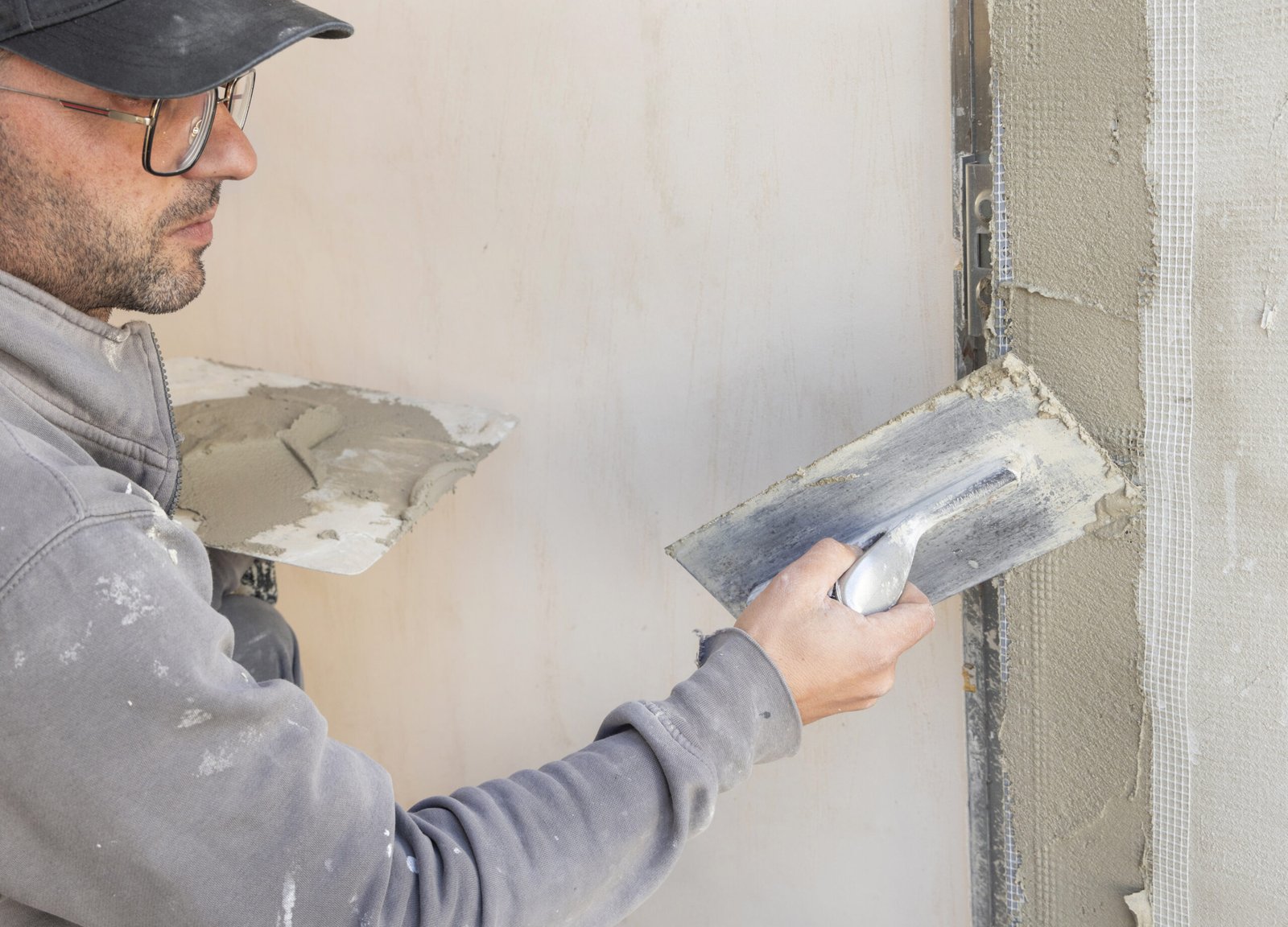 Craftsman uses a float trowel and cement to plaster a house wall. Young men with glasses in gray work clothing during construction or renovation process