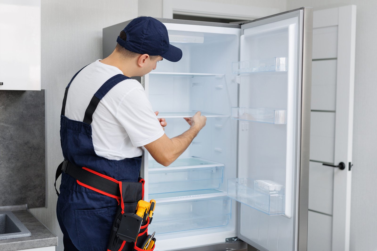 Man in a uniform with tools is repairing a refrigerator in the kitchen