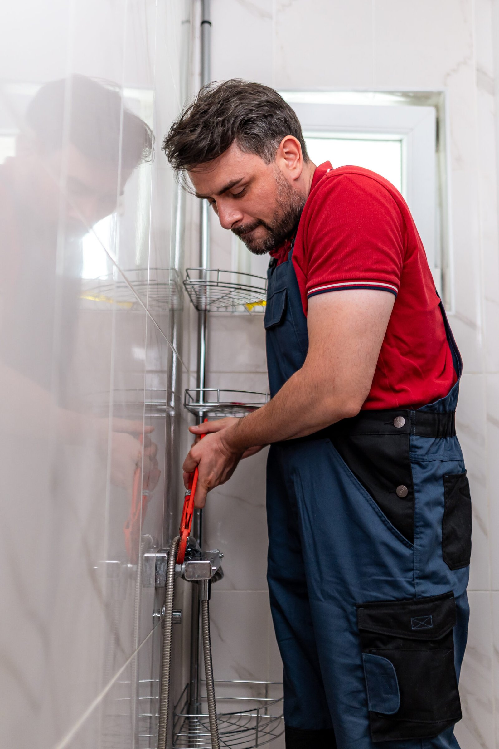 A man in a red shirt and blue overalls is fixing pipes in a bright bathroom. He uses tools and focuses on the task at hand.